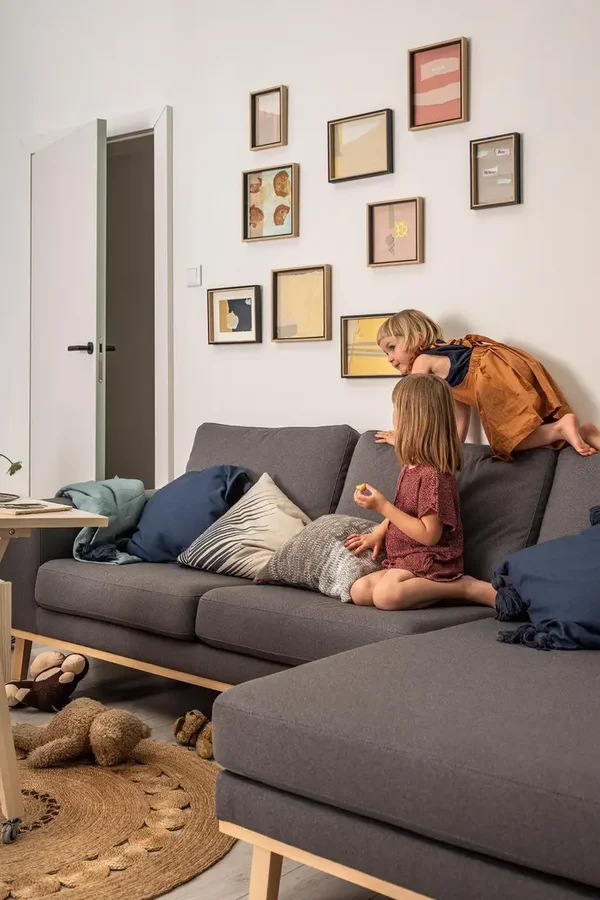  Jasne wnętrze z szarą sofą, bawiącymi się dziećmi i galerią rysunków na ścianie.